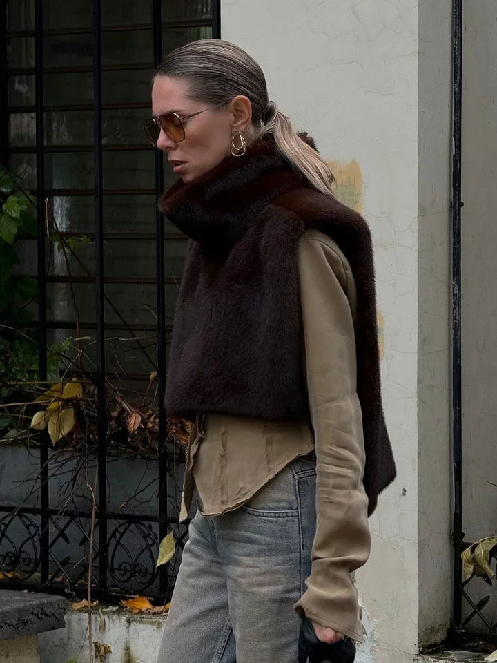 Kvinde iført brun pelsvest, beige skjorte og solbriller, går forbi et gitterhegn. Mode, efterårstøj, stilfuldt look, bymiljø.