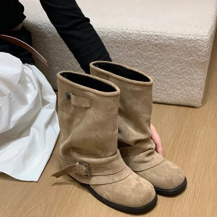 Beige ruskindsstøvler med spænde, placeret på trægulv ved siden af person i sort trøje og hvid nederdel. Stilfuldt fodtøj til efterårsmode.