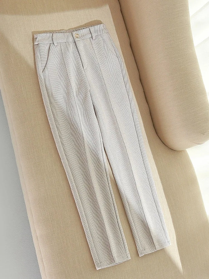 Beige bukser i hørblanding med høj talje og pressefolder, liggende på en lys sofa. Stilfuldt og moderne design til forår og sommermode.