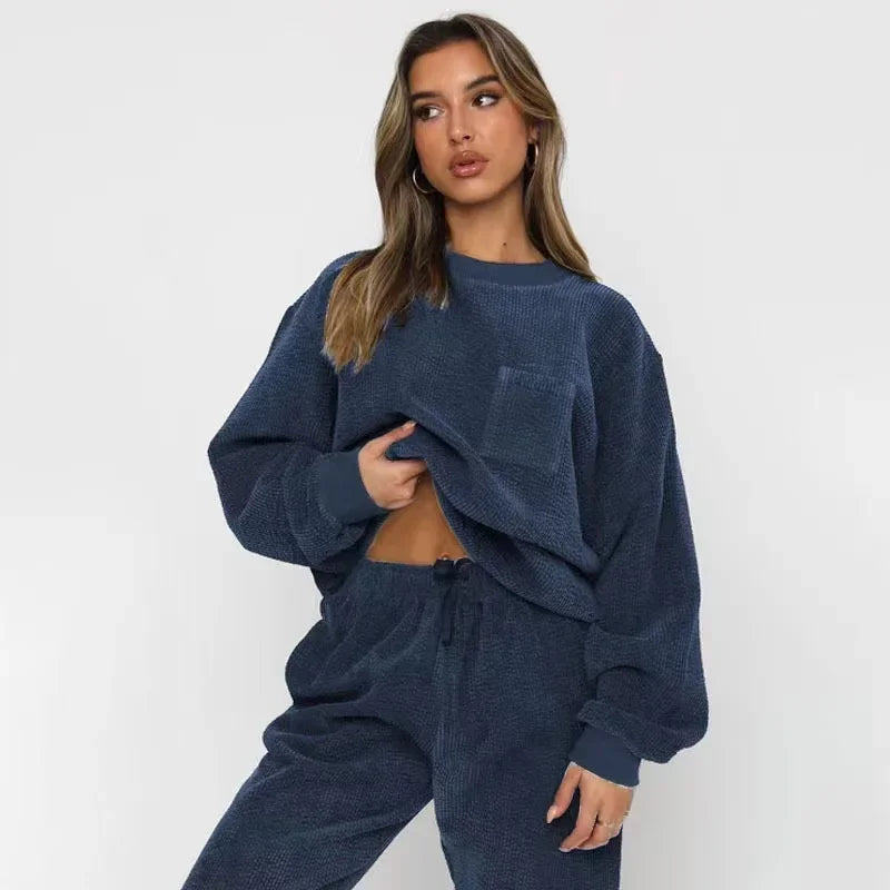 Kvinde iført blå, oversized sweatshirt og bukser, poserer mod hvid baggrund. Komfortabelt loungewear, moderne stil, afslappet mode.
