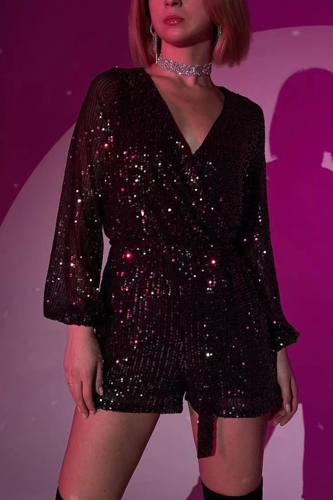 Kvinde i sort, glitrende jumpsuit med lange ærmer og V-udskæring, står mod en lilla baggrund. Perfekt til festmode og glamourøse events.