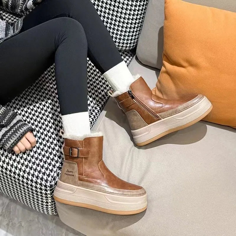 Brune læderstøvler med tykke såler og lynlås, båret af person i sorte leggings og hvide sokker, sidder på grå sofa med orange pude.