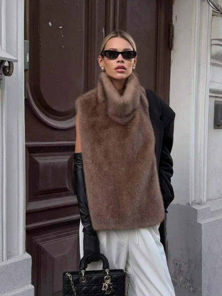 Kvinde i brun pelsvest, sorte solbriller og lange handsker, står foran en trædør. Hun bærer en sort taske og hvide bukser. Mode, vinterstil.