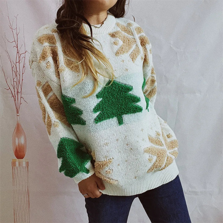 Kvinde iført hvid julesweater med grønne juletræer og beige snefnug, stående foran en lys baggrund. Perfekt til vintermode og juletema.