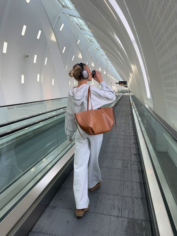 Kvinde i gråt joggingsæt med brun taske og hovedtelefoner går på rulletrappe i moderne lufthavn. Rejse, mode, komfort.