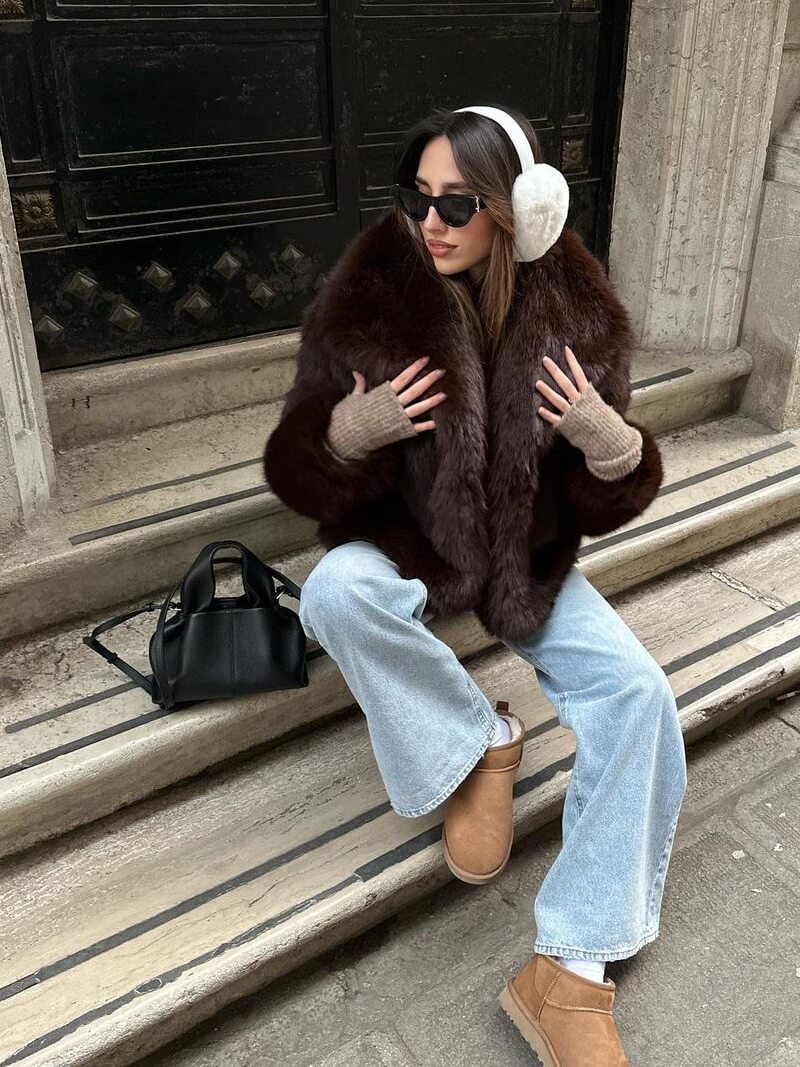 Kvinde i brun pelsjakke, lyseblå jeans og brune støvler sidder på trappe. Bærer solbriller og hvide ørevarmere. Sort taske ved siden af.