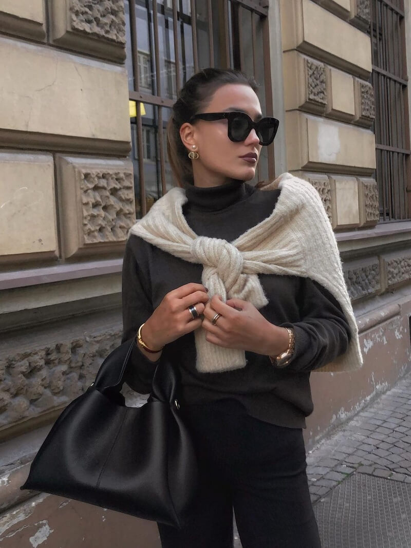 Kvinde i moderne efterårsoutfit med solbriller, sort taske og beige sweater over skuldrene, står foran en historisk bygning. Fashionabelt gadebillede.