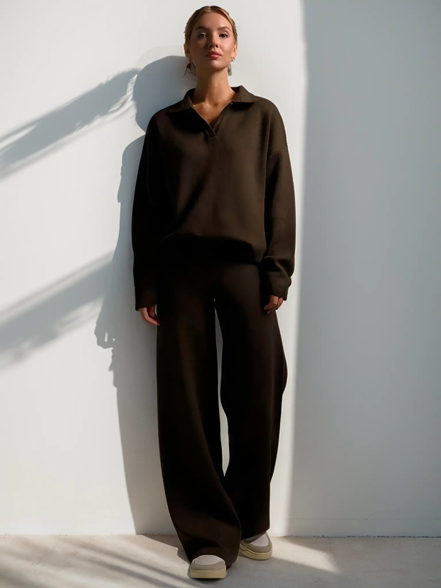 Kvinde iført mørkebrun, løstsiddende buksedragt står op ad en hvid væg. Minimalistisk mode, komfortabelt tøj, moderne stil.