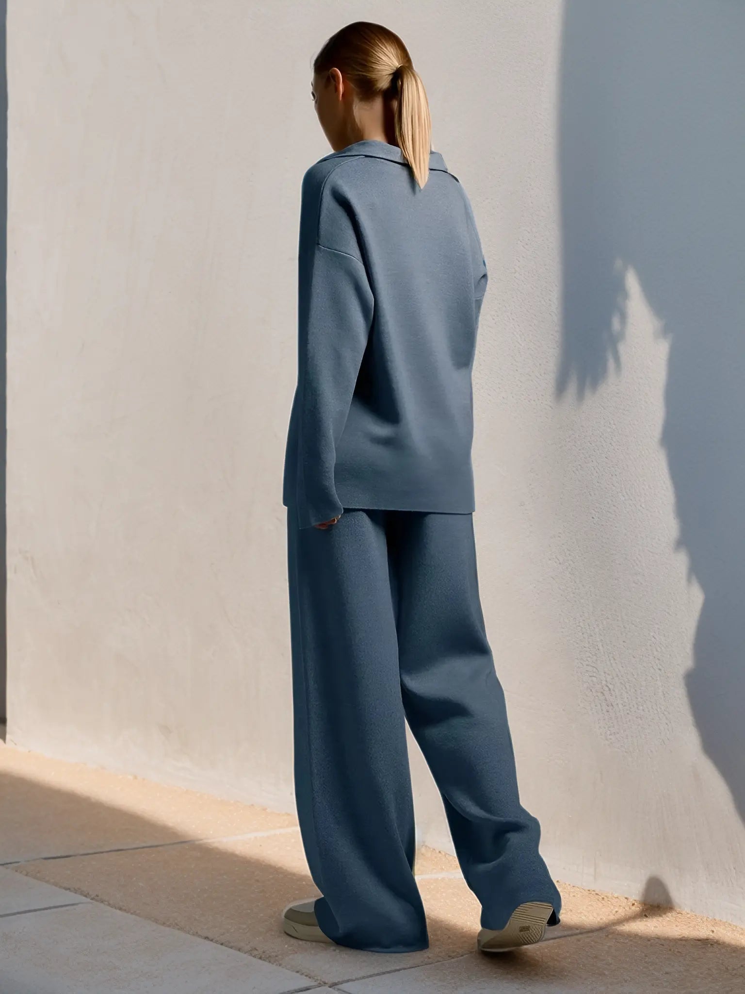 Kvinde iført blå, løstsiddende jakkesæt med bukser, står ved hvid væg. Moderne minimalistisk mode, elegant stil, afslappet look.