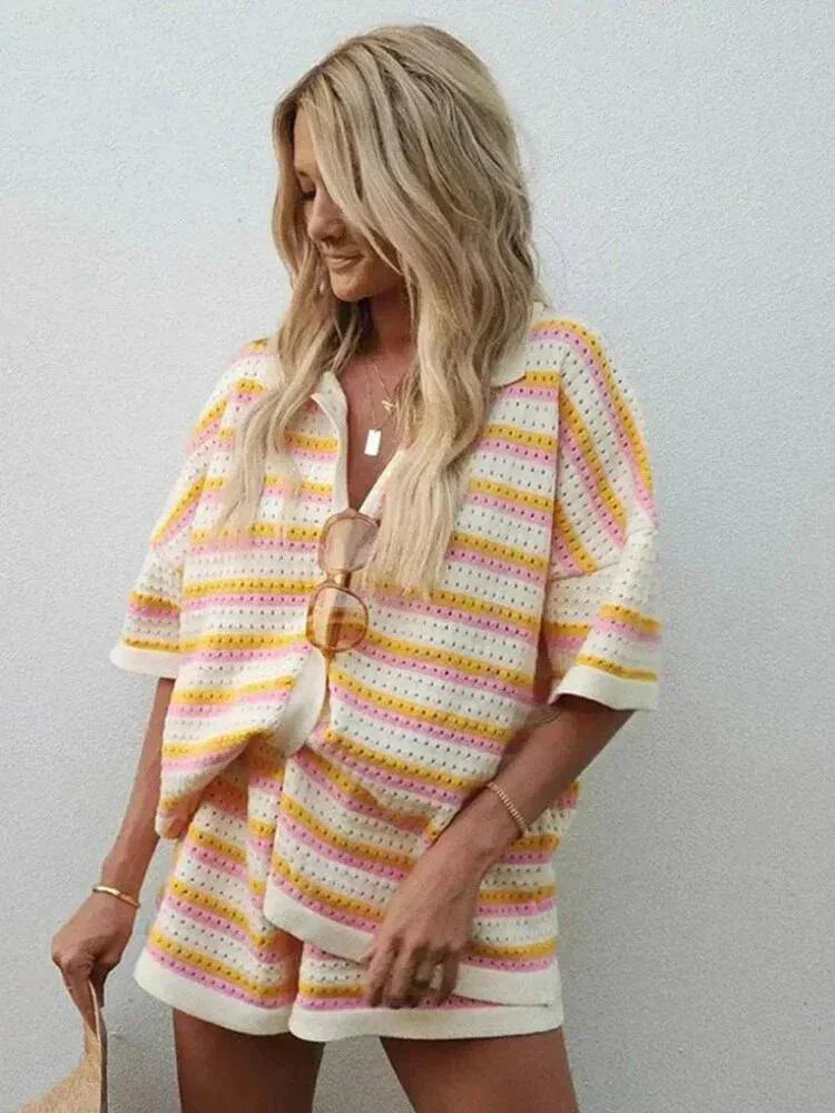 Blond kvinde i farverigt stribet sæt med kortærmet skjorte og shorts, poserer mod hvid væg. Sommermode, afslappet stil, trendy outfit.