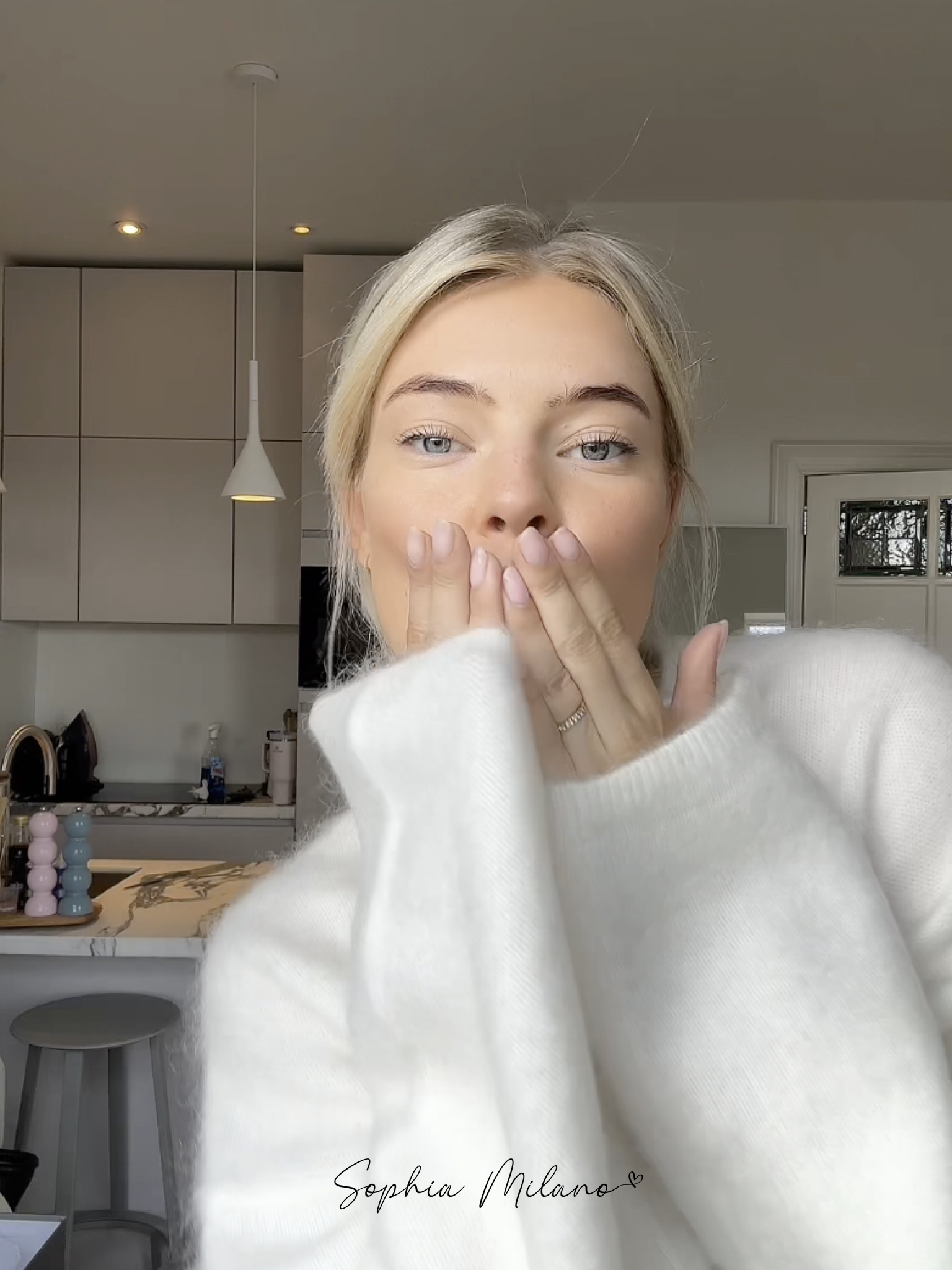 Blond kvinde i hvid sweater blæser et kys i moderne køkken. Skandinavisk interiør, minimalistisk design, naturligt lys.