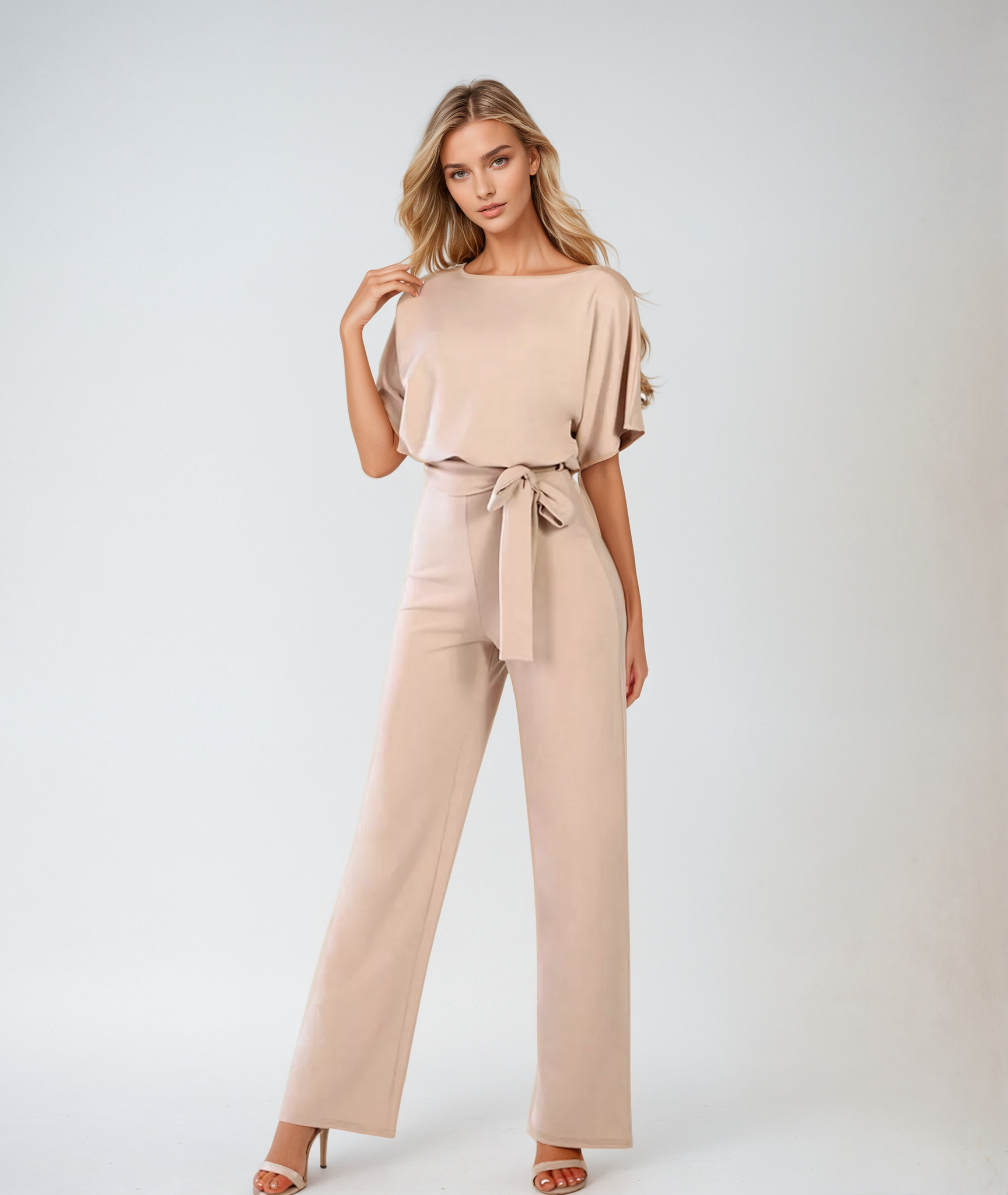 Blond kvinde i elegant beige jumpsuit med bælte, poserer mod hvid baggrund. Mode, stilfuldt tøj, moderne kvindemode, minimalistisk design.