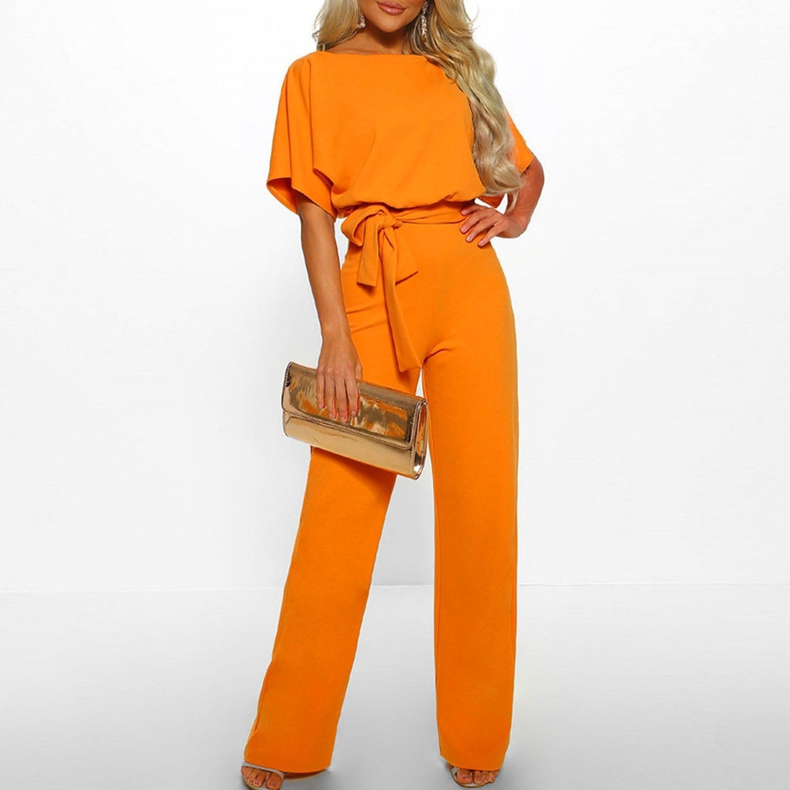 Kvinde iført orange buksedragt med bælte, holder en gylden clutch. Moderne mode, elegant stil, perfekt til fest eller aftenbegivenheder.