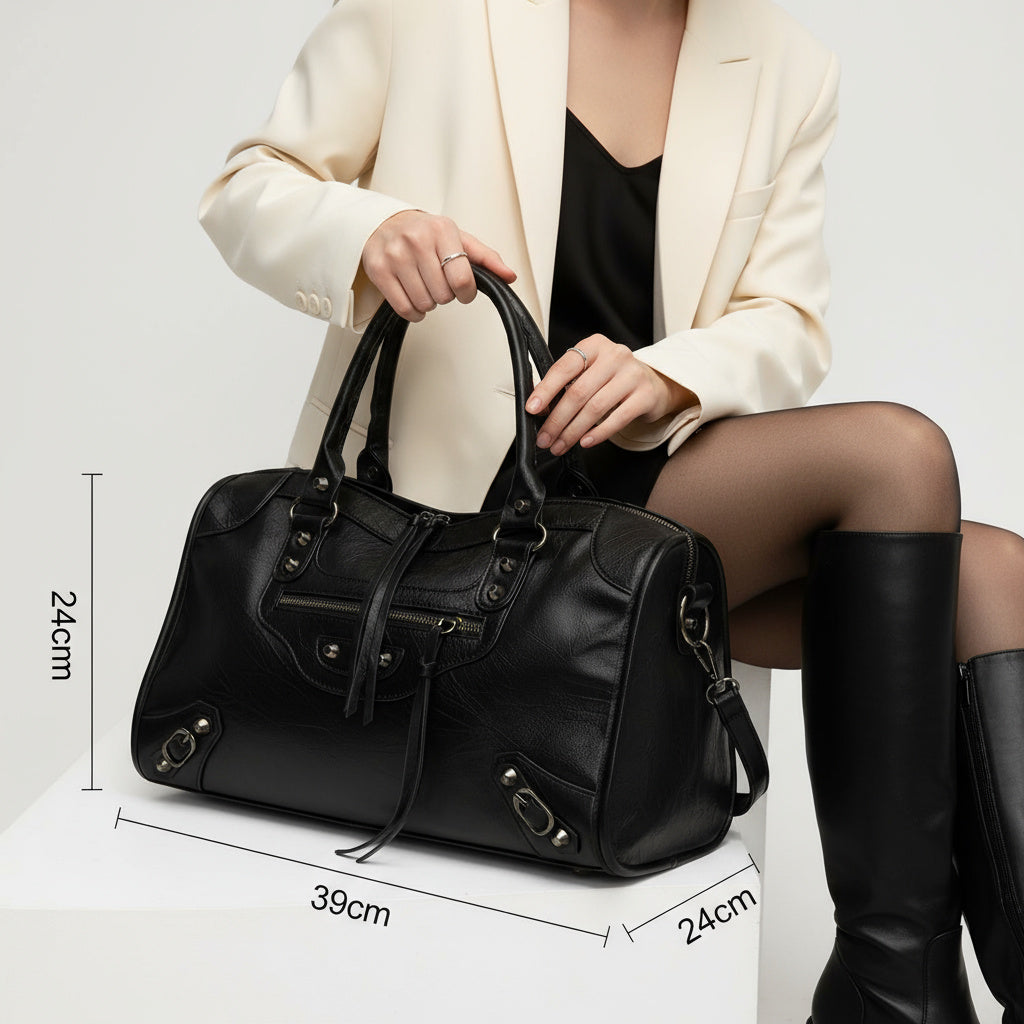 Kvinde i hvid blazer med sort lædertaske, 39x24 cm, sidder på hvid kasse. Tasken har lynlåse og håndtag. Mode, tilbehør, stilfuld taske.