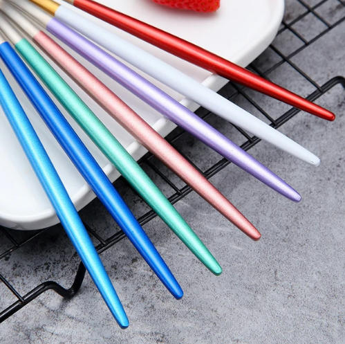 Farverige metalspisepinde på hvid tallerken, grå baggrund. Moderne design, genanvendelige, miljøvenlige. Perfekt til asiatisk madlavning.