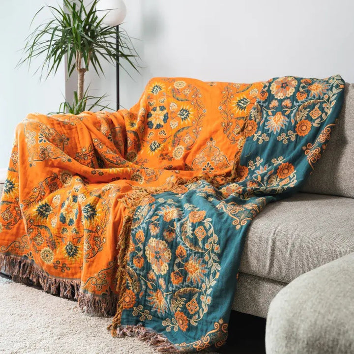 Orange og blå blomstret tæppe på grå sofa i stue med grøn plante. Farverigt indretningstilbehør, boheme stil, hyggelig boligindretning.