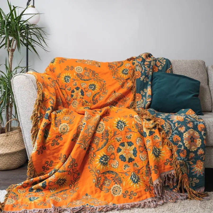 Orange og blå blomstret tæppe på grå sofa med grøn pude, stueindretning, boheme stil, hyggelig atmosfære, moderne hjem, plante i kurv ved siden af.