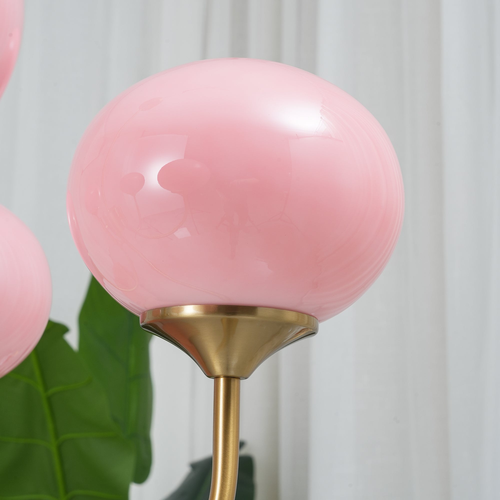 Lyserød kugleformet lampe med gylden base foran hvidt gardin og grøn plante. Moderne indretning, stilfuld belysning, elegant design.