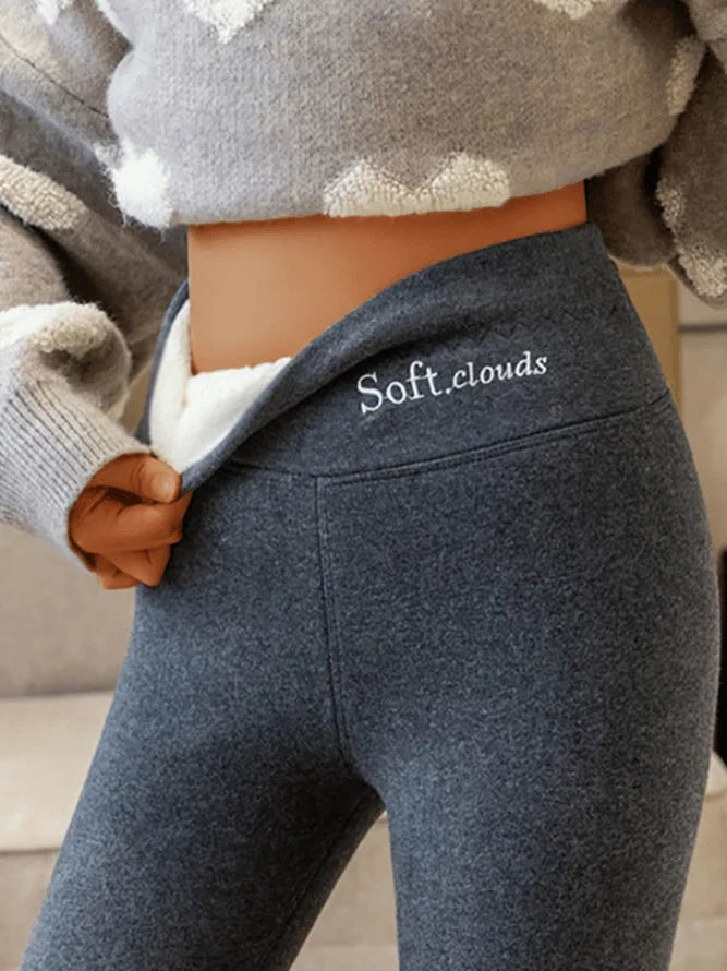 Kvinde iført grå leggings med "Soft.clouds" logo, trækker i linningen. Komfortabelt vintertøj, blødt stof, moderne design.