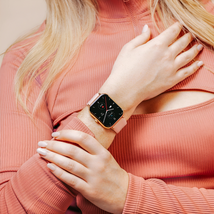 Kvinde iført lyserød smartwatch med firkantet urskive og silikonerem, matcher lyserød trøje. Moderne teknologi, mode, wearable tech.