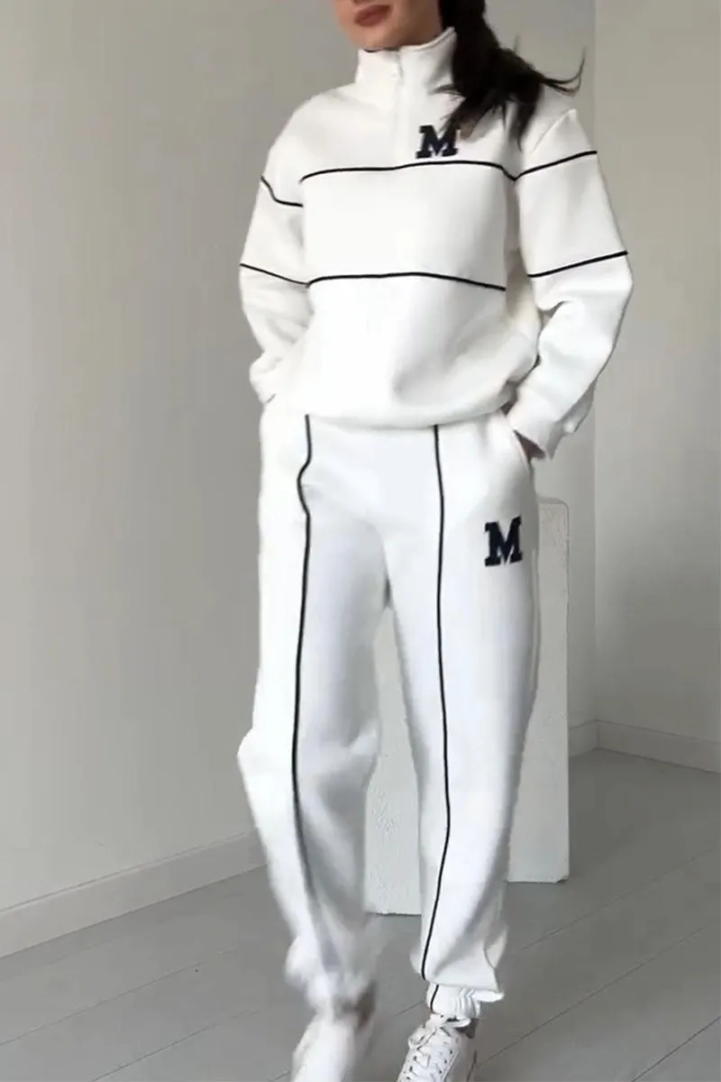 Kvinde iført hvid træningsdragt med sorte striber og "M" logo, står i minimalistisk rum. Sportstøj, mode, komfortabelt outfit, casual stil.