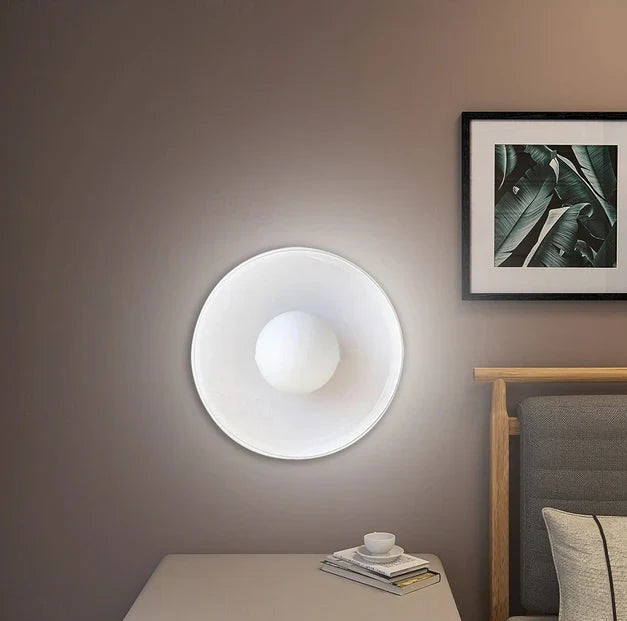 Væglampe i moderne design ved siden af seng, oplyser værelse med blødt lys. Minimalistisk indretning med kunstværk og natbord.