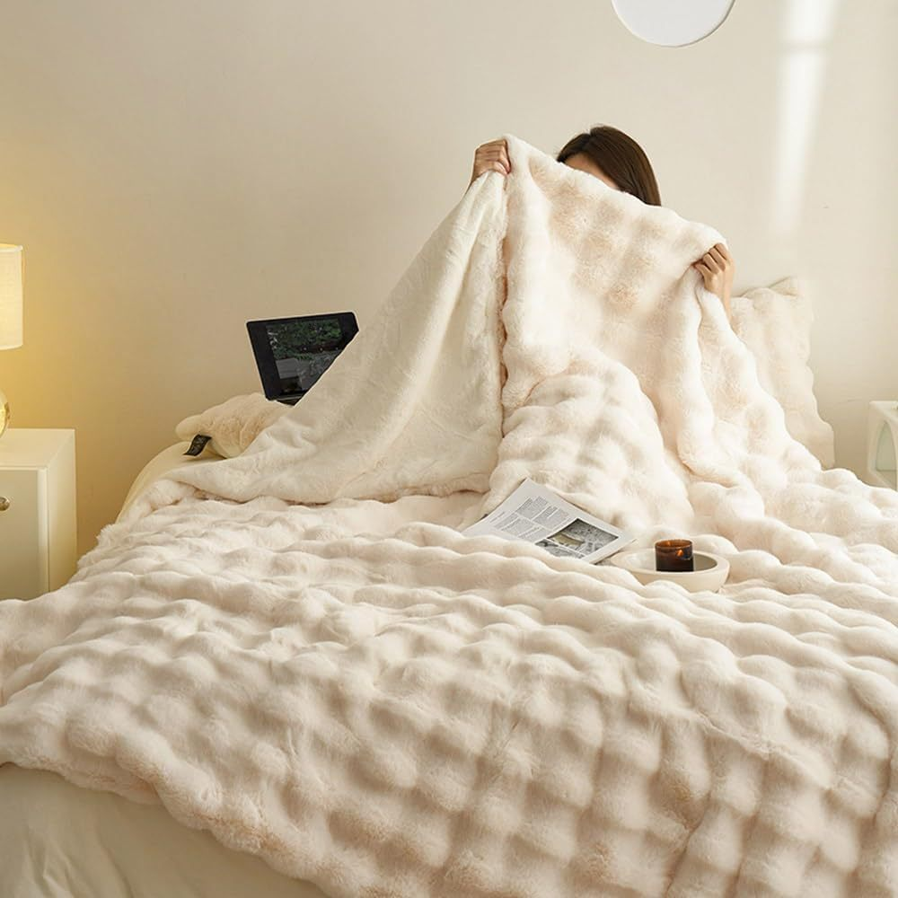 Kvinde gemmer sig under blød, hvid plysplaid på seng med laptop og magasin. Hygge, komfort, afslapning i soveværelse.