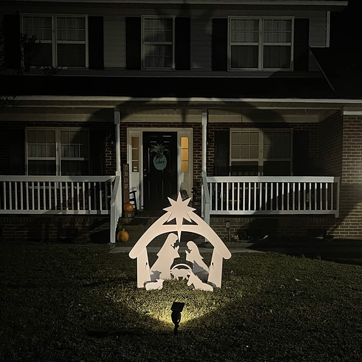 Hvid julekrybbe dekoration oplyst i nat foran et hus med veranda. Julestemning, udendørs pynt, natbelysning, juledekoration.