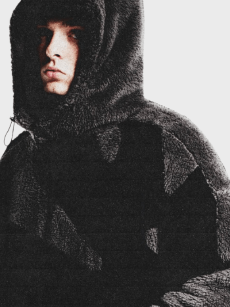 Ung mand iført en sort, fluffy hættetrøje med hætte, ser direkte mod kameraet. Vintermode, moderne herretøj, stilfuldt look.