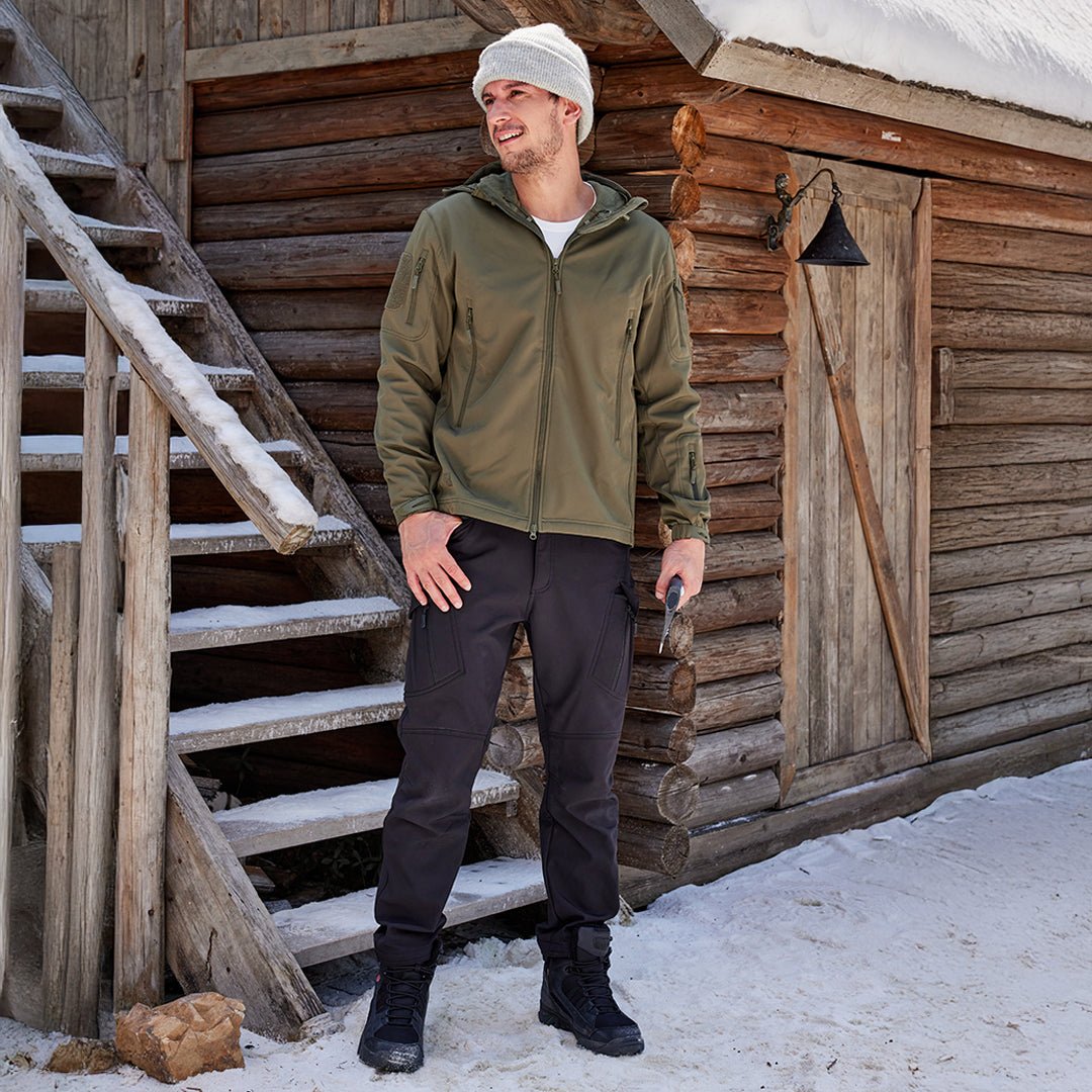 Mand i vintertøj står foran træhytte med sne, iført grøn jakke, sorte bukser og støvler. Vintermode, udendørs, skandinavisk stil.