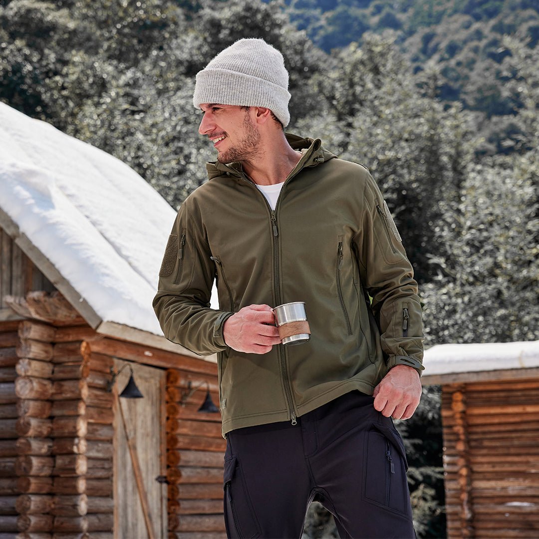 Mand i grøn jakke og hvid hue står foran en træhytte i sneklædt landskab, holder en kop. Vintermode, udendørs, afslapning, natur.