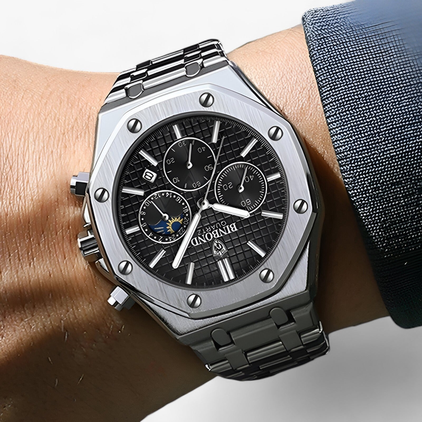 Sølv armbåndsur med sort urskive, kronograffunktioner og månefaseindikator. Elegant herreur i rustfrit stål, moderne design.