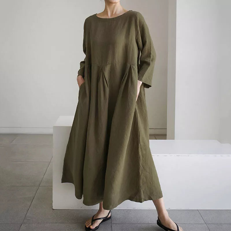 Kvinde iført en løs, olivengrøn maxikjole med lange ærmer og lommer, stående i minimalistisk rum. Komfortabelt og stilfuldt hverdagslook.