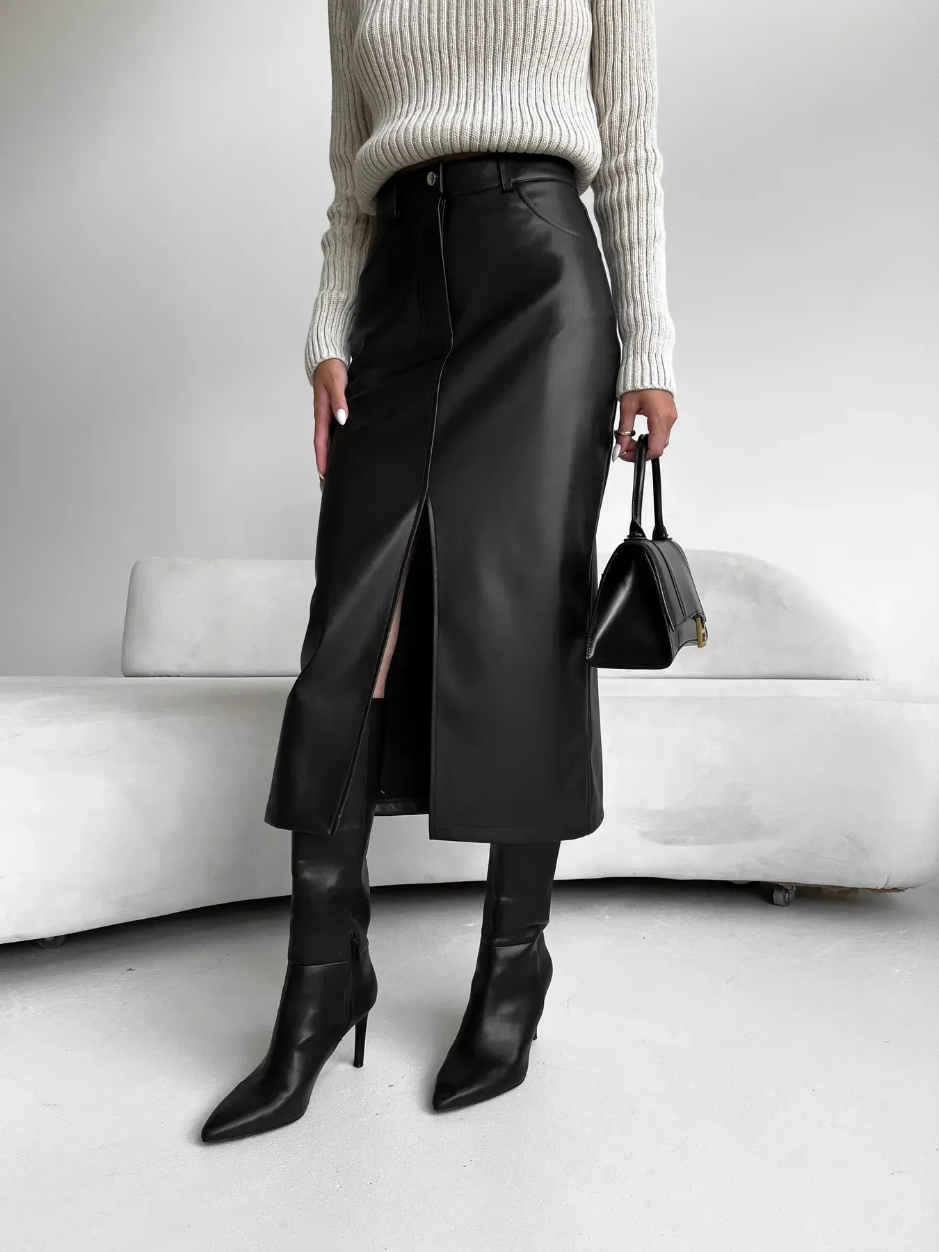 Kvinde iført sort lædernederdel med slids, beige striktrøje og sorte støvler. Holder en sort håndtaske. Moderne mode, stilfuldt outfit.