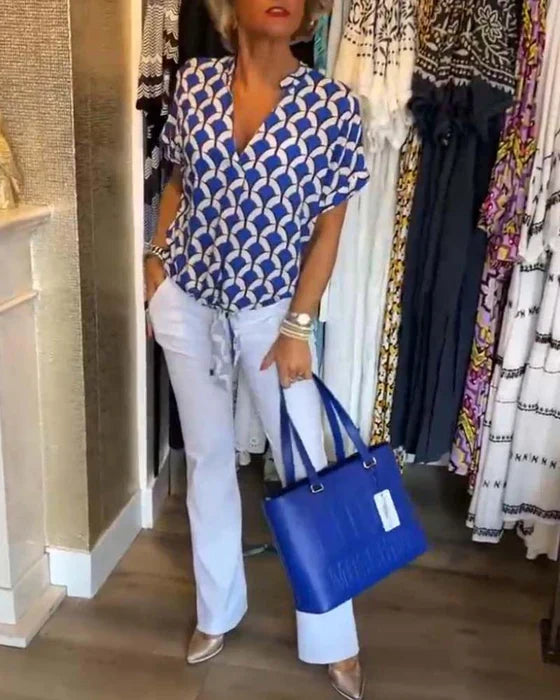 Kvinde i blå-hvid mønstret bluse og hvide bukser, holder en blå taske. Står i tøjbutik med farverige kjoler i baggrunden. Mode, shopping, stil.