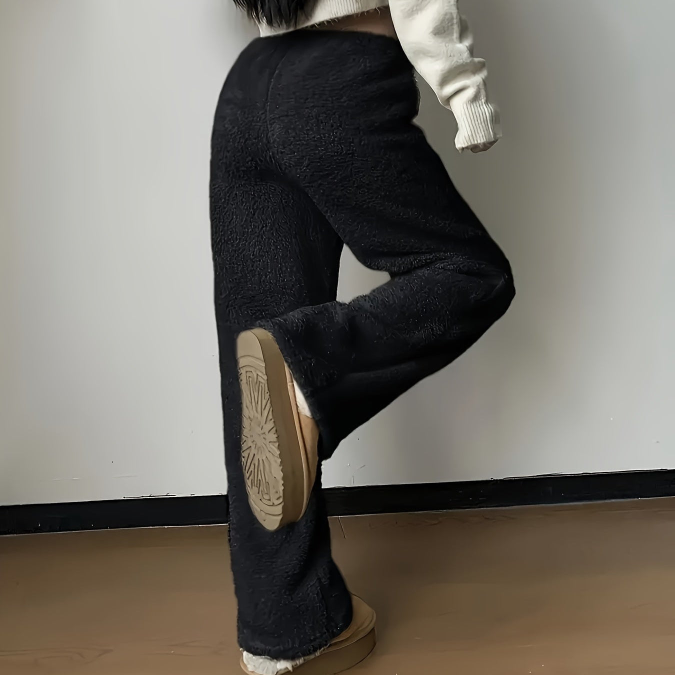 Kvinde iført sorte fleecebukser og beige sweater, står på trægulv. Fokus på komfortabelt vintertøj, mode, og afslappet stil.