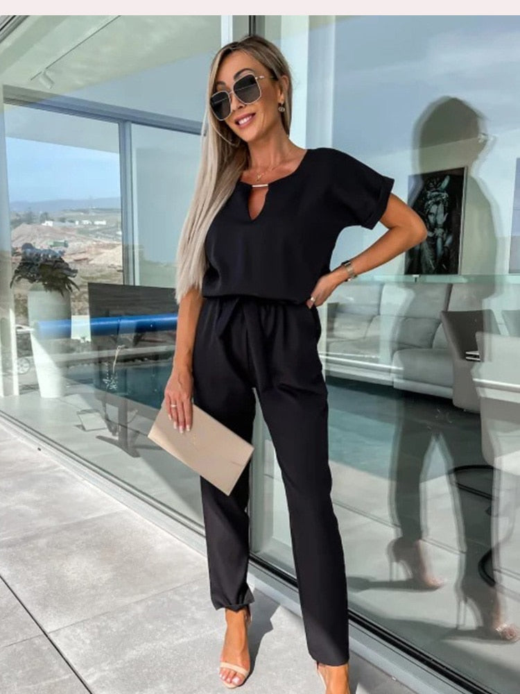 Kvinde i sort jumpsuit med solbriller og beige clutch foran moderne glasbygning. Stilfuldt outfit, mode, sommerlook, udendørs.